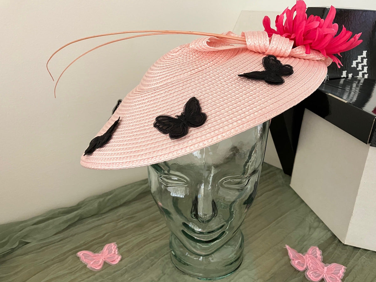 A light pink woven fascinator hat with black butterfly appliqués and a bright pink feather flower, displayed on a clear glass mannequin head.
