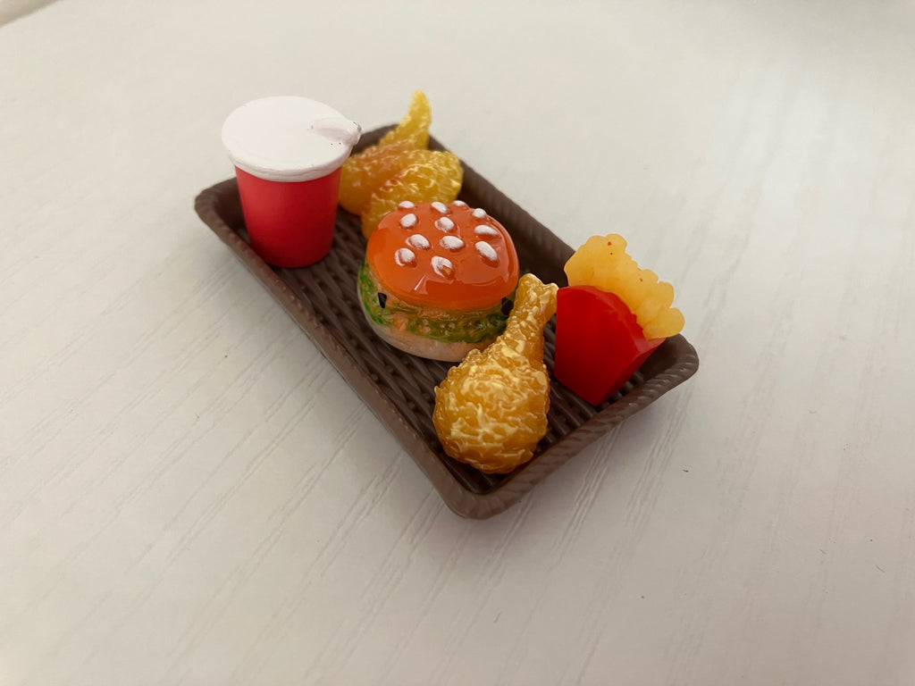 A miniature toy food set on a small brown tray, featuring a hamburger with a red bun, a red cup with a white lid, two golden french fries, and a golden chicken leg, displayed on a light wood surface.
