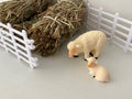 Two small plastic toy sheep figurines, one standing and one lying down, displayed on a light surface with miniature white fence panels and hay bales in the background.