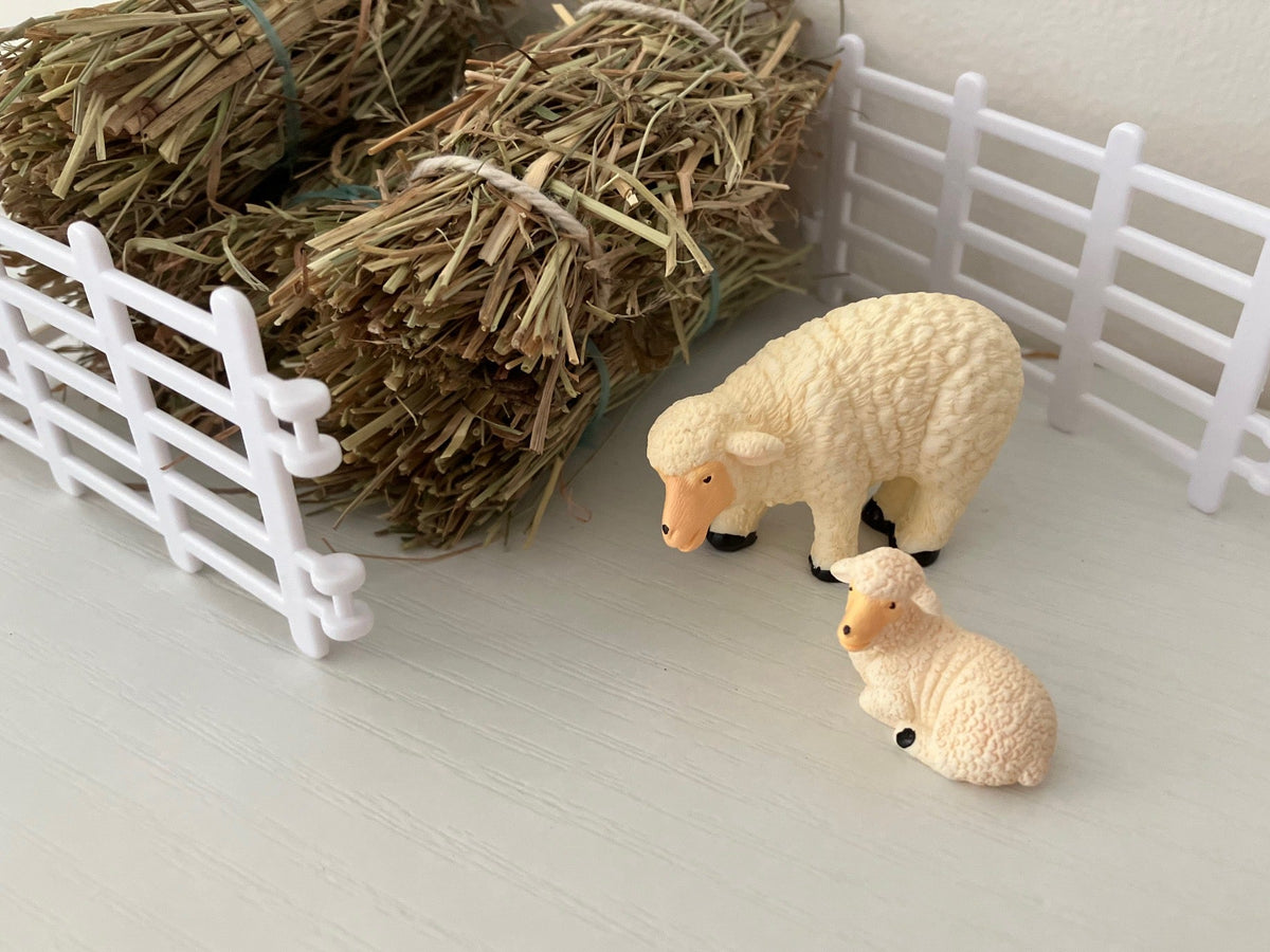 Two small plastic toy sheep figurines, one standing and one lying down, displayed on a light surface with miniature white fence panels and hay bales in the background.