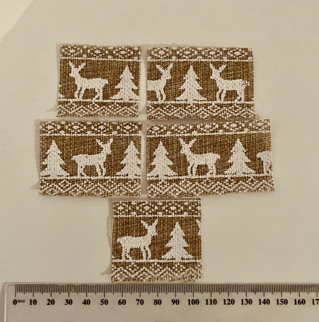 Five small rectangular fabric patches with a brown burlap-like background and white embroidered holiday motifs of deer, trees, and geometric borders, arranged on a light beige surface with a ruler at the bottom for scale.
