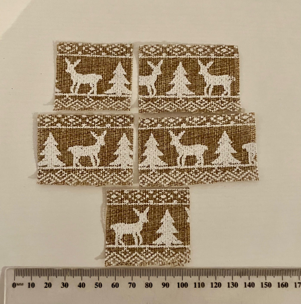 Five small rectangular fabric patches with a brown burlap-like background and white embroidered holiday motifs of deer, trees, and geometric borders, arranged on a light beige surface with a ruler at the bottom for scale.