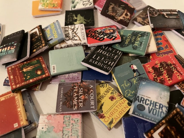 A pile of miniature book-shaped magnets or small book covers spread out on a light surface, showing many different book designs and titles in assorted colors.