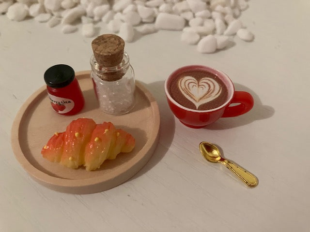 A miniature coffee-themed miniature scene featuring a small red ceramic cup of latte with heart-shaped foam art, a small red jar, a miniature gold spoon, and a pastry on a wooden tray.