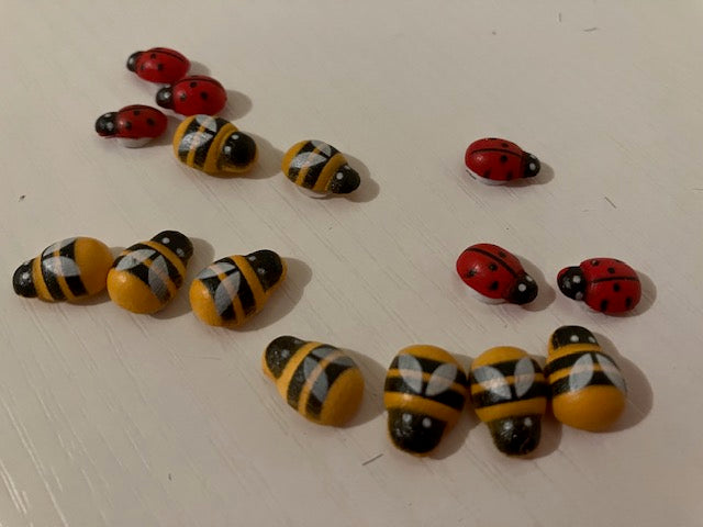 A set of decorative beads shaped like bumblebees and ladybugs scattered on a light off-white surface. The beads feature yellow and black stripes on the bodies and red and black spots on the heads and shells.