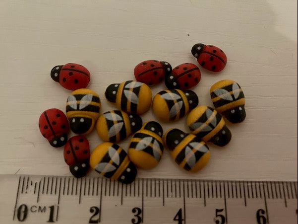 A group of decorative beads shaped like ladybugs and bees, displayed on a light surface with a ruler at the bottom for scale. The beads feature red ladybugs with black spots, yellow and black striped bees, and white ladybugs with black spots.