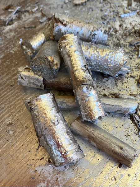 A pile of natural birch wood fire starter logs with silver-colored bark, resting on a wooden surface with sawdust.