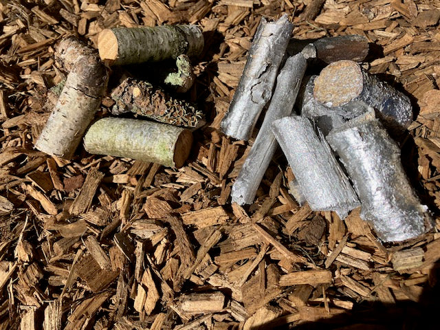 A pile of small natural wood pieces, bark pieces, and a pinecone scattered on a bed of brown wood chips.