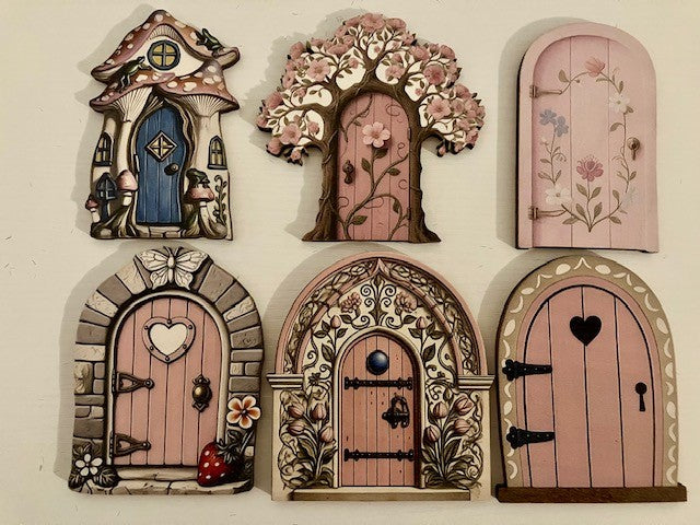 Six decorative miniature fairy doors and tree arches displayed on a light beige wall. The set includes whimsical designs such as a mushroom house with a blue door, a tree with pink blossoms, a pink floral arch, a stone-brick arch with a heart-shaped door, an ornate floral arch, and a pink heart-shaped door.